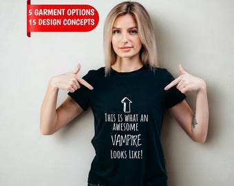 May include: A black short-sleeved t-shirt featuring a white upward-pointing arrow graphic and the text "THIS IS WHAT AN AWESOME VAMPIRE LOOKS LIKE!" printed on the front. The shirt is paired with dark gray denim jeans.