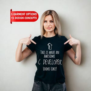 May include: A black short-sleeved t-shirt featuring a white graphic of an upward-pointing arrow and the text: THIS IS WHAT AN AWESOME C DEVELOPER LOOKS LIKE! A red banner in the upper left corner displays the text: 5 GARMENT OPTIONS 15 DESIGN CONCEPTS.