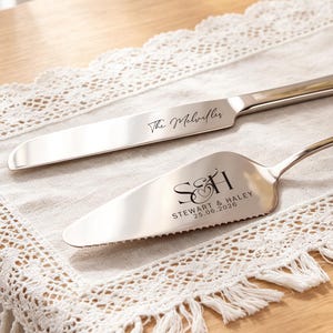May include: A silver-toned cake knife and server set featuring custom engravings. The knife is inscribed with "The Melvilles" and the server displays the initials "S&H" above the text "STEWART & HALEY" and the date "25.06.2026" in a dark font.