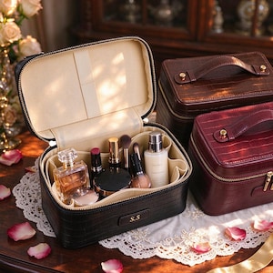 Puede incluir: Tres estuches cosméticos rectangulares con textura de cocodrilo, con cremalleras doradas y asas superiores. Un estuche negro está abierto, mostrando un interior beige con varios productos de belleza, mientras que dos estuches cerrados son marrón y burdeos.