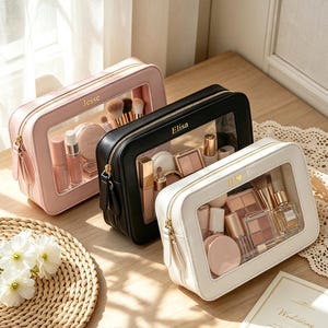 Puede incluir: Tres neceseres rectangulares en rosa claro, negro y blanco, cada uno con un panel frontal transparente, cremallera dorada y texto personalizado. El neceser rosa tiene el nombre Jesse, el negro Elisa y el blanco muestra unas iniciales.