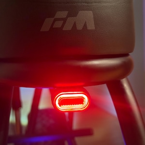 Puede incluir: Taburete de cuero negro con una lámpara roja. El taburete tiene un asiento acolchado con un logotipo en relieve. La luz roja tiene forma ovalada y emite un brillo intenso. Las patas del taburete son oscuras y curvas.