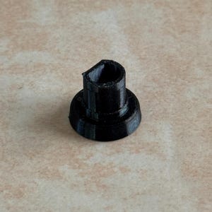 May include: A black, 3D-printed bottle stopper. The stopper has a cylindrical body with a flared base and a notched top. The object is made of a matte black material and sits on a light brown surface.