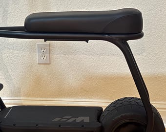 May include: A matte black electric scooter featuring a long, cushioned seat mounted on a tubular metal frame. The scooter has a wide, flat deck with a logo, a single rear tire with a deep tread pattern, and a small black foot peg near the front.