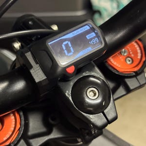 Può includere: Primo piano di un display digitale nero sul manubrio di una bici elettrica Sur-Ron. Il display mostra "0" e "499 km/h". Un pulsante rosso è visibile sotto lo schermo. Il manubrio e i componenti circostanti sono neri.