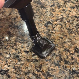 May include: Black metal stand with a square base and a rounded top. The stand is attached to a black metal rod. The base has a textured, layered appearance. The stand is on a speckled brown and tan surface.