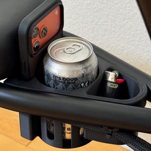May include: A black plastic accessory tray mounted on a frame, featuring a dedicated slot for a smartphone, a circular holder containing a silver beverage can with the text IT'S GOOD SINCE 1898, and a small compartment holding a black BIC lighter.