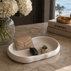 May include: A white, oval-shaped ceramic catch-all tray with a smooth, organic rim. Inside the tray sits a beige quilted leather card holder with a gold logo, a black car key fob, and a gold-tone wristwatch with a black dial and metal link band.