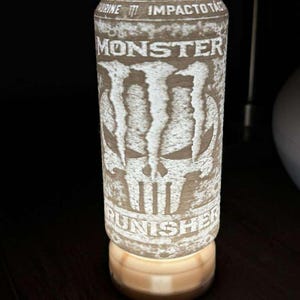 Puede incluir: L&aacute;mpara cil&iacute;ndrica con dise&ntilde;o de lata de bebida energ&eacute;tica Monster. La lata presenta el logotipo de Monster y un gr&aacute;fico de calavera, con la palabra "PUNISHER" en la parte inferior. La l&aacute;mpara emite un brillo c&aacute;lido y suave desde su base.