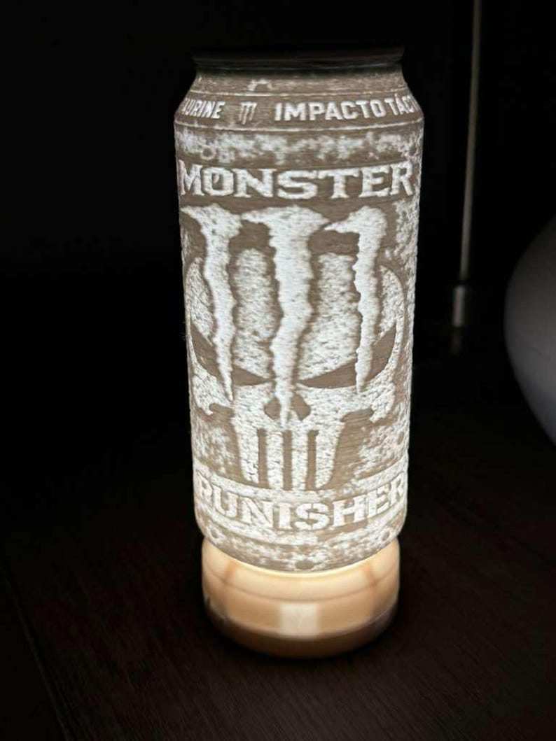 Puede incluir: L&aacute;mpara cil&iacute;ndrica con dise&ntilde;o de lata de bebida energ&eacute;tica Monster. La lata presenta el logotipo de Monster y un gr&aacute;fico de calavera, con la palabra "PUNISHER" en la parte inferior. La l&aacute;mpara emite un brillo c&aacute;lido y suave desde su base.