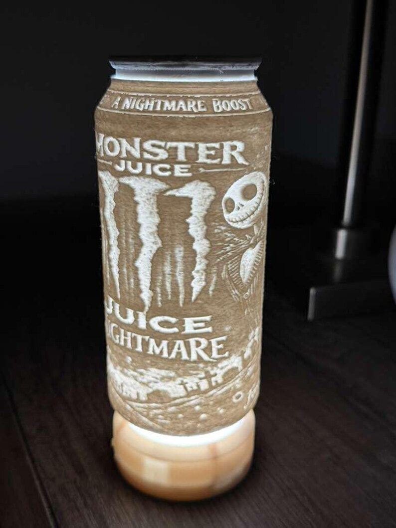 Puede incluir: L&aacute;mpara cil&iacute;ndrica marr&oacute;n con las palabras "Monster Juice" y "Juice Nightmare" grabadas en la superficie. La l&aacute;mpara presenta un dise&ntilde;o de Jack Skellington de The Nightmare Before Christmas. La l&aacute;mpara se apoya en una base de madera y emite un brillo suave.