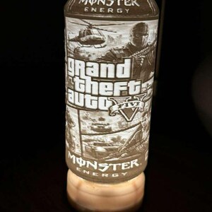 Puede incluir: Una lata de bebida Monster Energy con un dise&ntilde;o de Grand Theft Auto V. La lata presenta un gr&aacute;fico detallado de un helic&oacute;ptero, una persona sosteniendo un arma y el logotipo del juego. La lata est&aacute; iluminada desde abajo.