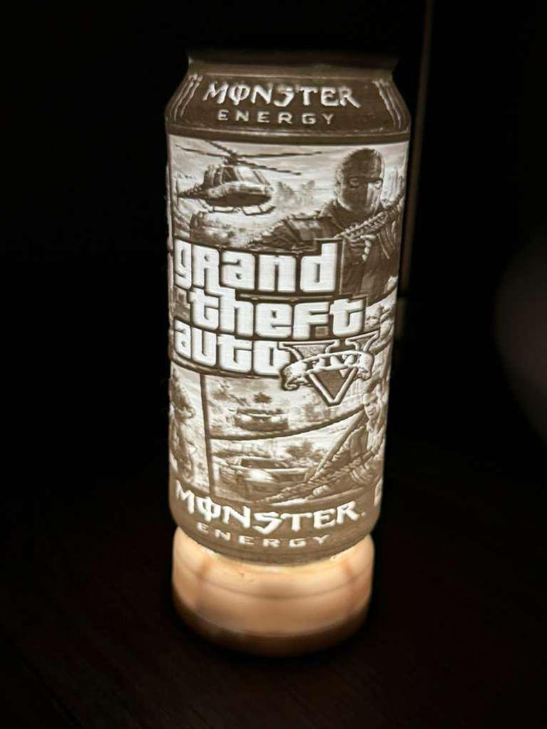 Puede incluir: Una lata de bebida Monster Energy con un dise&ntilde;o de Grand Theft Auto V. La lata presenta un gr&aacute;fico detallado de un helic&oacute;ptero, una persona sosteniendo un arma y el logotipo del juego. La lata est&aacute; iluminada desde abajo.