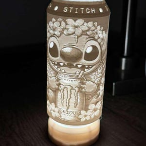 Puede incluir: L&aacute;mpara decorativa con forma de lata, con un relieve detallado del personaje Stitch. La l&aacute;mpara tiene la palabra "STITCH" en la parte superior y el logotipo de Monster Energy en la lata. La base de la l&aacute;mpara emite una luz c&aacute;lida.