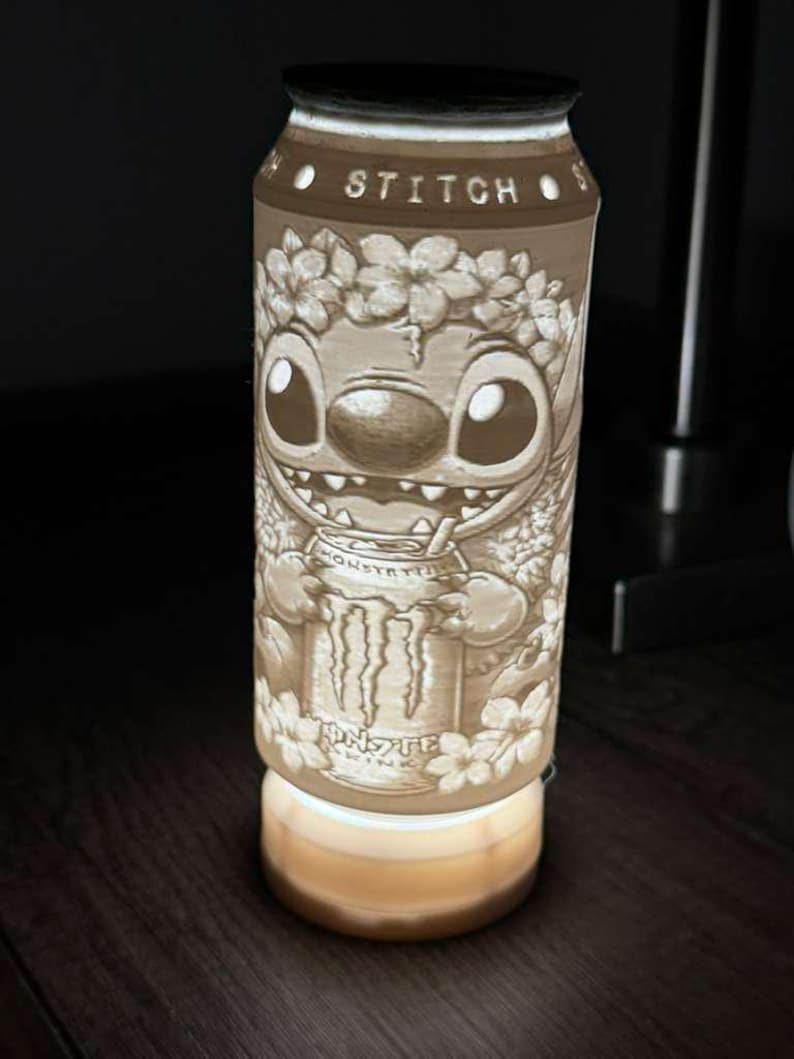 Puede incluir: L&aacute;mpara decorativa con forma de lata, con un relieve detallado del personaje Stitch. La l&aacute;mpara tiene la palabra "STITCH" en la parte superior y el logotipo de Monster Energy en la lata. La base de la l&aacute;mpara emite una luz c&aacute;lida.