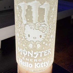 Puede incluir: L&aacute;mpara cil&iacute;ndrica iluminada con un dise&ntilde;o de Hello Kitty y Monster Energy. La l&aacute;mpara es blanca con detalles grabados, incluyendo las palabras "HELLO KITTY" y "MONSTER ENERGY". La base es de color c&aacute;lido.