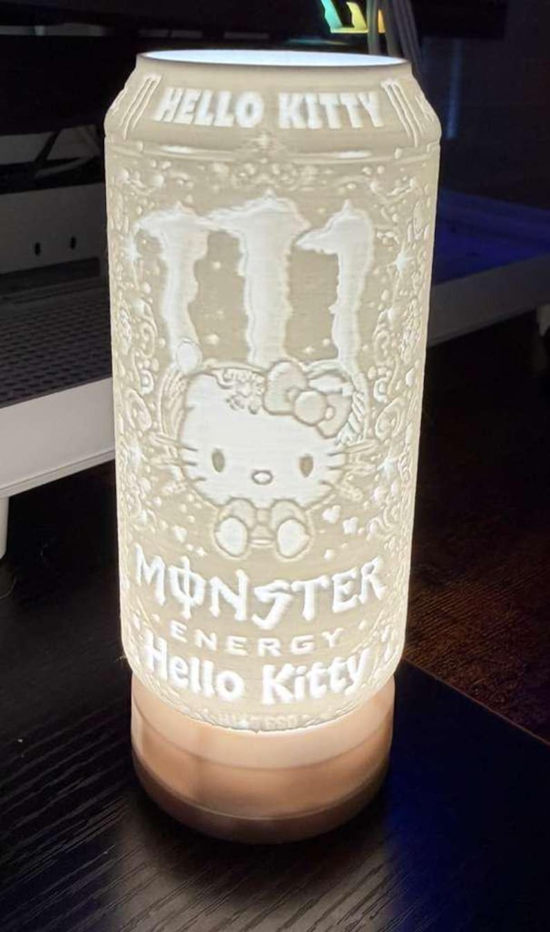 Puede incluir: L&aacute;mpara cil&iacute;ndrica iluminada con un dise&ntilde;o de Hello Kitty y Monster Energy. La l&aacute;mpara es blanca con detalles grabados, incluyendo las palabras "HELLO KITTY" y "MONSTER ENERGY". La base es de color c&aacute;lido.
