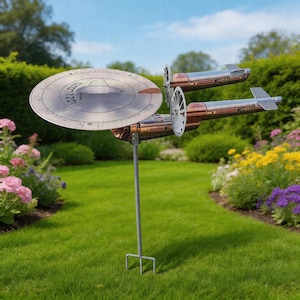 Puede incluir: Una estaca de jardín de metal con diseño de nave espacial. La nave plateada y cobriza tiene una sección frontal circular con el texto "NCC-1701" y dos secciones laterales. La estaca está en un jardín con césped y flores.