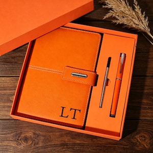 May include: Orange leather journal and pen set in a gift box. The journal has a strap closure and the initials "LT" embossed on the cover. A silver pen and an orange pen are included.