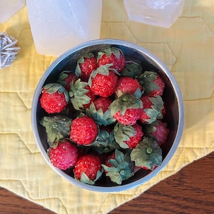 May include: A metal bowl filled with artificial strawberries. The strawberries are bright red with green leaves. The bowl sits on a yellow quilted surface, with white crystal formations in the background.