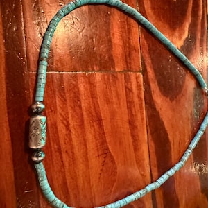 May include: Turquoise beaded necklace with a silver centerpiece. The necklace features small, rectangular turquoise beads strung together. The centerpiece is a silver cylinder with a starburst design. The necklace is displayed on a wooden surface.