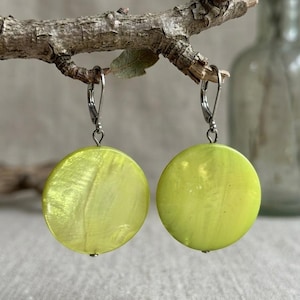 May include: A pair of lime green, round earrings with silver-colored lever-back closures. The earrings are hanging from a small, textured tree branch. The background is a neutral, light gray color.