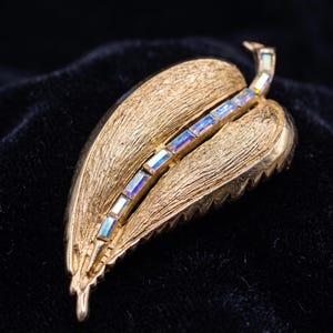 May include: A gold-toned leaf-shaped brooch with a textured surface. The brooch features a central line of iridescent rectangular crystals. The leaf design has a detailed vein pattern, adding to its vintage appeal.