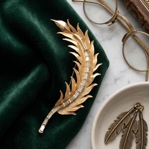 May include: A gold-toned feather brooch with clear rectangular crystals, resting on a dark green velvet surface. Beside it are gold-framed eyeglasses, a small dish with two feather-shaped earrings, and a vintage book.