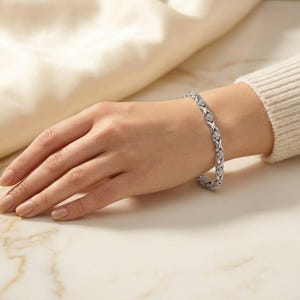 May include: A silver-toned bracelet featuring a repeating X-shaped link design. Each X-shaped link is accented with small, clear, sparkling stones set along the surface. The bracelet has a secure clasp mechanism and a polished metallic finish.