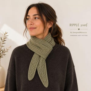 Op de afbeelding: Een groene gebreide sjaal, om de nek gewikkeld, met de tekst "RIPPLE scarf" en "By DesignedMorocco HANDMADE KNIT PATTERN". De sjaal heeft een geribbelde textuur en is gemaakt van een zacht materiaal. Het model draagt een donkerbruine trui.