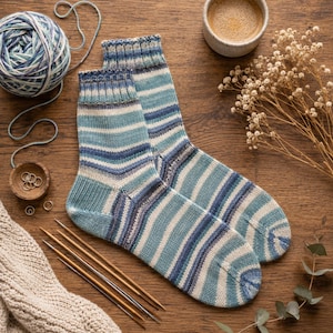 Peut inclure: Paire de chaussettes tricotées à la main avec des rayures horizontales bleues, turquoise et blanches. Les chaussettes sont posées sur une surface en bois, avec des aiguilles à tricoter, de la laine et une tasse de café.
