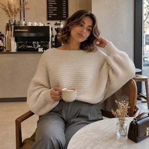 以下が含まれることがあります： クリーム色のセーターとグレーのパンツを着た人が、コーヒーカップを持っています。カフェに座っており、背景にはコーヒーメーカーとメニューが見えます。メニューにはコーヒーと価格が記載されています。