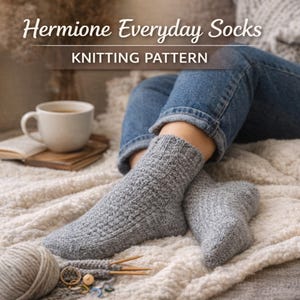 Könnte beinhalten: Graue Strickstrümpfe, Teil des Strickmusters "Hermione Everyday Socks", werden gezeigt. Das Bild zeigt einen Wollknäuel, Stricknadeln und eine Tasse Kaffee, was auf ein gemütliches, handgemachtes Thema hindeutet.