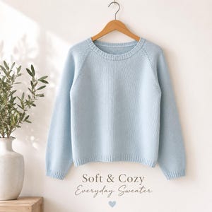 Könnte beinhalten: Ein hellblauer Pullover mit Rundhalsausschnitt und langen Ärmeln hängt an einem Holzbügel. Der Pullover scheint aus einem weichen Material zu sein. Der Text "Soft & Cozy Everyday Sweater" wird unter dem Pullover angezeigt.