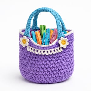 May include: A handmade crochet basket in a vibrant purple color, featuring a blue handle and decorative white and yellow flower accents. The basket is filled with colorful clothespins. A charming and functional storage solution.