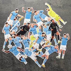 以下が含まれることがあります： 水色と黒のユニフォームを着たサッカー選手のイラストが描かれたステッカーのコレクション。ステッカーは、サッカーボールを持っているものも含め、さまざまな選手のアクションポーズを描いています。背景は濃いグレーの表面です。
