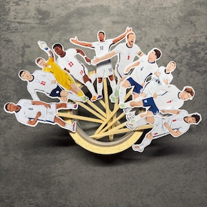 May include: A collection of paper cutouts depicting soccer players in white and yellow uniforms, each attached to a wooden toothpick. The players are shown in various action poses, featuring jersey numbers like 23, 10, 9, 4, 21, 8, and 6.