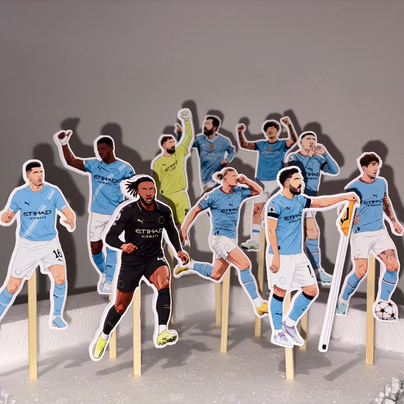 May include: A collection of soccer player cutouts on wooden sticks. The players are wearing light blue and white uniforms, with some in black. The cutouts are arranged on a white surface against a gray background. A soccer ball is also included.