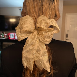 Pode incluir: Um grande laço decorativo para cabelo feito de tecido cor de champanhe com um padrão de glitter dourado metálico apresentando espirais e motivos de folhas. O laço tem bordas com arame e duas caudas longas que pendem, preso a um rabo de cavalo.