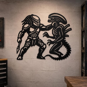 May include: Black metal wall art depicting a Predator and an Alien in a battle pose. The Predator is holding a spear, and the Alien has its arms outstretched. The artwork is mounted on a gray concrete wall.