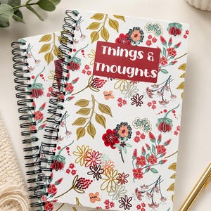 May include: Two spiral-bound notebooks with a white floral pattern featuring red, gold, and teal flowers and leaves. A red sticker on the front of one notebook reads "Things & Thoughts". A gold pen rests on the surface.