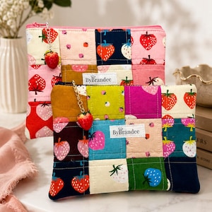May include: Two rectangular quilted pouches featuring a patchwork design of various colored squares with strawberry prints. Each pouch has a pink or dark blue zipper closure with a small strawberry charm attached and a white label reading ByBrandee.