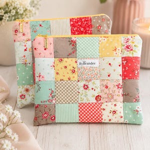 May include: Two rectangular zippered pouches made of quilted fabric with a patchwork design featuring various floral, gingham, and polka dot patterns in shades of red, yellow, green, and brown. Each pouch has a gold zipper and a ByBrandee label.
