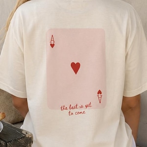 Könnte beinhalten: Ein cremefarbenes T-Shirt mit kurzen Ärmeln und einem großen Aufdruck auf der Rückseite, der ein Herz-Ass zeigt. Das Kartendesign enthält den Buchstaben A, ein rotes Herzsymbol und den kursiven Text "the best is yet to come" in roter Schrift.