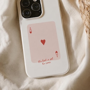 Può includere: Una cover per smartphone bianca con una grafica rettangolare rosa chiaro progettata per assomigliare a una carta da gioco asso di cuori. La grafica include simboli di cuori rossi e il testo "the best is yet to come" in corsivo rosso.