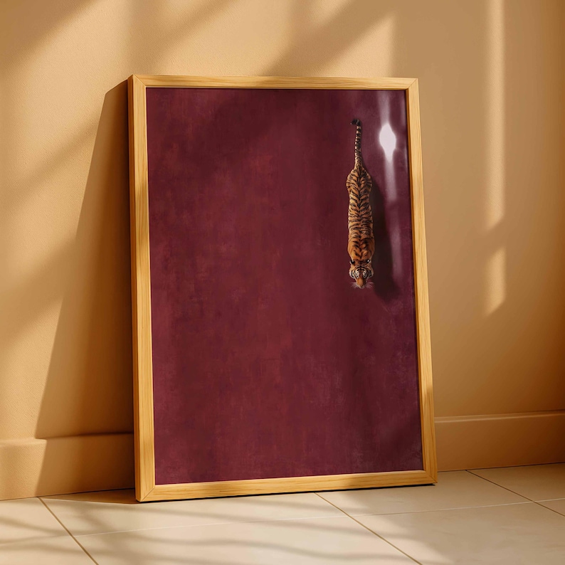 May include: A rectangular wall art print in a light wood frame featuring a tiger viewed from above, positioned on the right side against a solid deep burgundy background. The tiger has orange and black stripes and is facing downward.