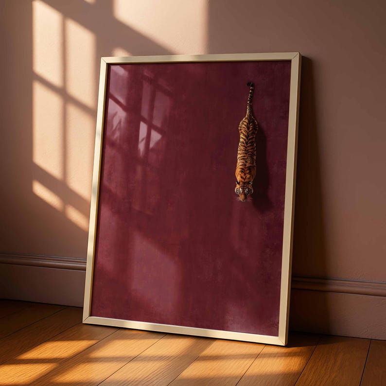 May include: A rectangular wall art piece in a thin gold frame featuring a deep burgundy background with a single tiger depicted in a downward-facing position. The tiger has orange and black stripes and is positioned in the upper right quadrant.