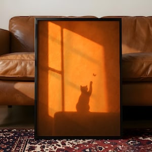 May include: A rectangular framed wall art print featuring a silhouette of a cat reaching toward a small butterfly. The image is dominated by warm orange and golden light, creating a window-like shadow effect across the entire composition.
