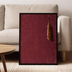 May include: A rectangular wall art piece featuring a deep burgundy velvet background enclosed in a thin black frame. A small, three-dimensional tiger figurine is attached to the right side of the velvet surface, positioned vertically with its head facing downward.