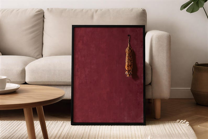 May include: A rectangular wall art piece featuring a deep burgundy velvet background enclosed in a thin black frame. A small, three-dimensional tiger figurine is attached to the right side of the velvet surface, positioned vertically with its head facing downward.